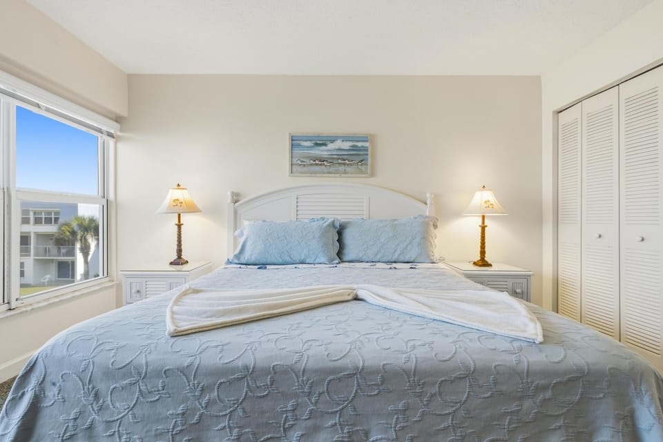 King bed with pale blue quilt and pillows, flanked by white nightstands and golden lamps, near a window view.
