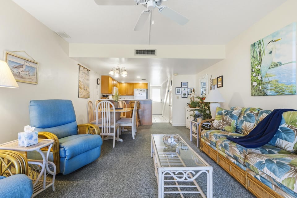 Living room with tropical-patterned sofa, blue recliner, glass-top rattan coffee table, and ceiling fan overhead.