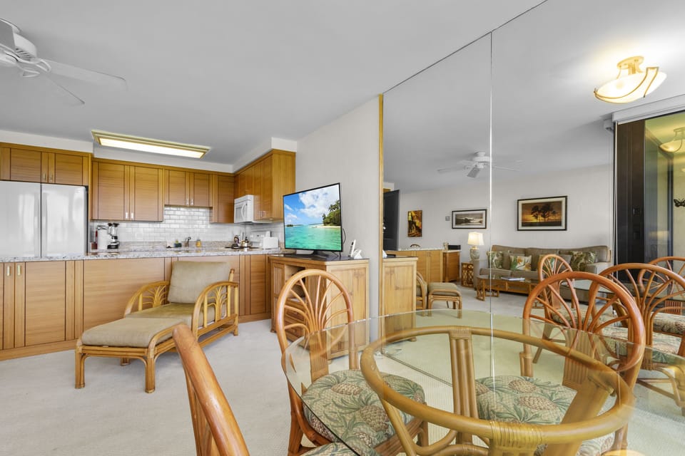 Gather around the elegant glass dining table, where tropical breezes flow through this open-concept space. Warm wood cabinetry and island seating create an inviting hub for memorable meals and laughter.