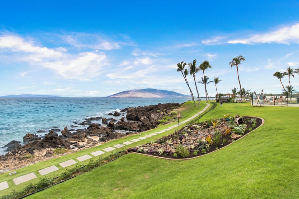 Oceanfront property features manicured grounds bordered by volcanic rock and palm trees, with a distant mountain visible across the water under clear blue skies.