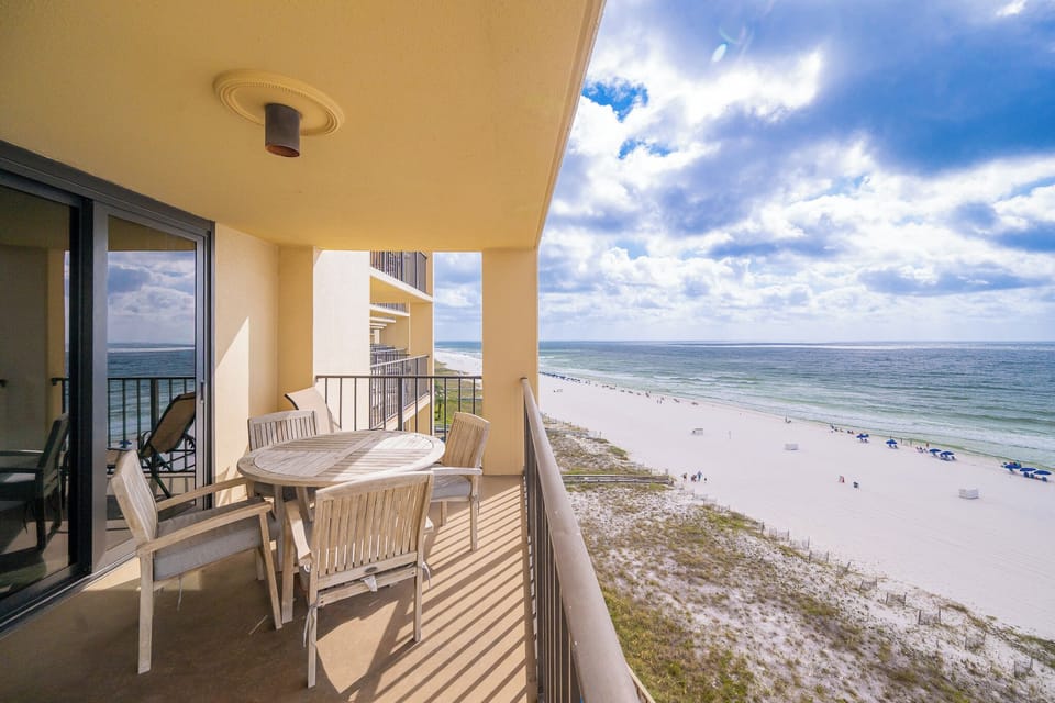 The balcony features a round wooden table with chairs, offering a view of the beach and gentle waves in the distance