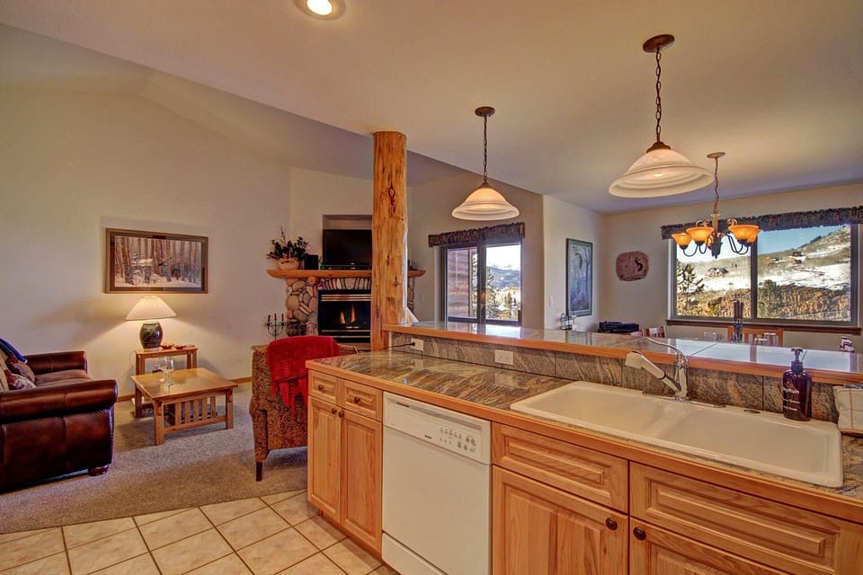 A cozy open-plan living room and kitchen area with wooden cabinetry, modern appliances, a leather sofa, a stone fireplace, large windows, and mountain views.
