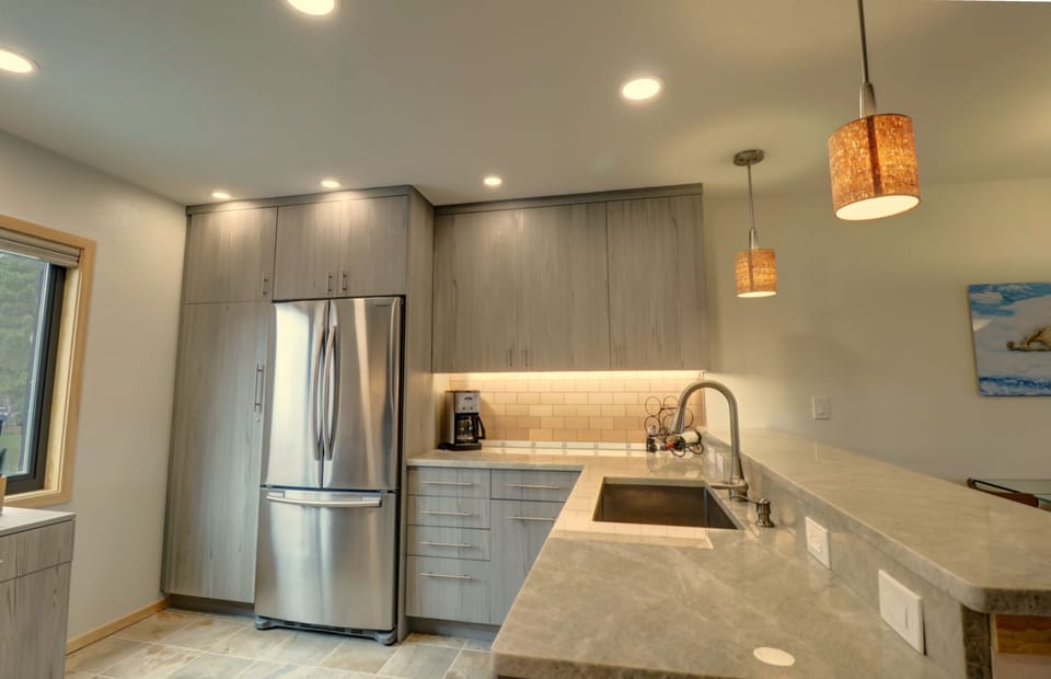 Modern kitchen with stainless steel refrigerator, large sink, grey cabinets, tiled backsplash, and marble countertops. Pendant lights hang over the counter. A window is seen to the left.