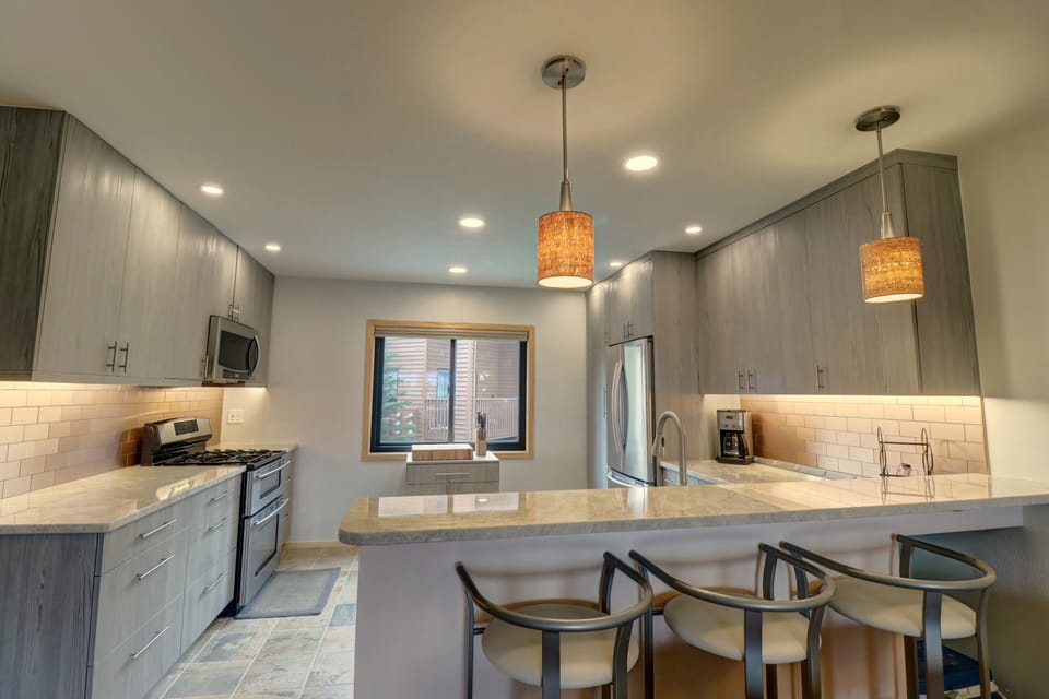 Modern kitchen with grey cabinets, stainless steel appliances, a central island with bar seating, pendant lights, and a window over the sink.