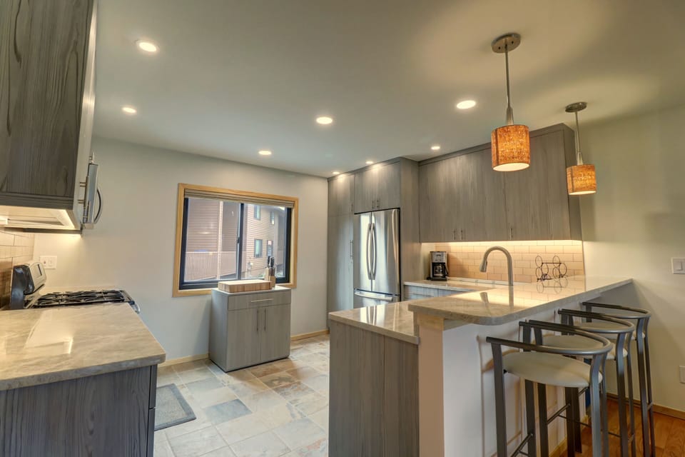 A modern kitchen with gray cabinets, stainless steel appliances, pendant lighting, and a breakfast bar with stools. The room is well-lit with large windows and recessed ceiling lights.