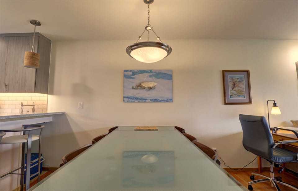 A minimalist dining area with a large glass-top table, chairs, a hanging light fixture, and a wall with two pieces of artwork. A small home office setup with a desk and chair is visible on the right.