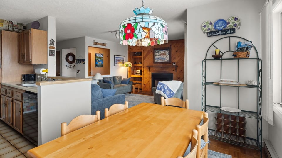 A cozy living room and dining area with a wooden table, chairs, blue couch, armchair, fireplace, and colorful stained glass light fixture. A kitchen with wooden cabinets is visible on the left.