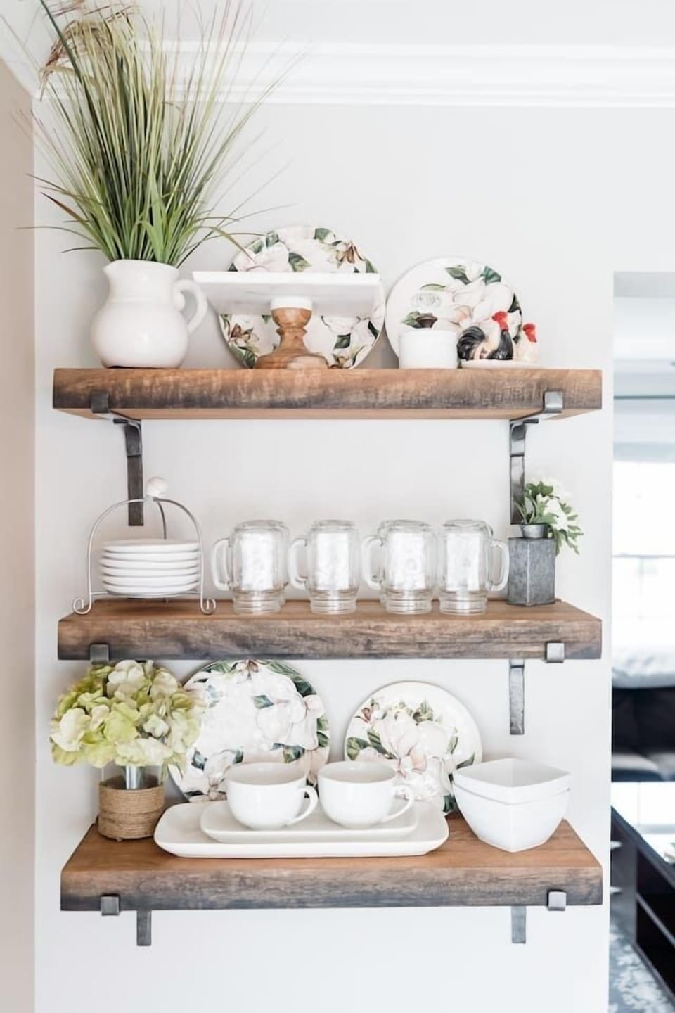 These charming rustic floating shelves are fully stocked with essentials, including mason jar mugs, decorative floral plates, and clean white bowls. Everything is neatly organized and easily accessible for your dining needs.