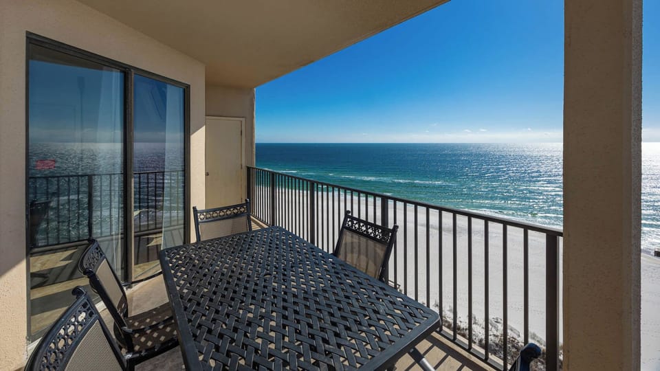 A spacious balcony features a black metal table and chairs, offering a breathtaking view of the beach and shimmering water
