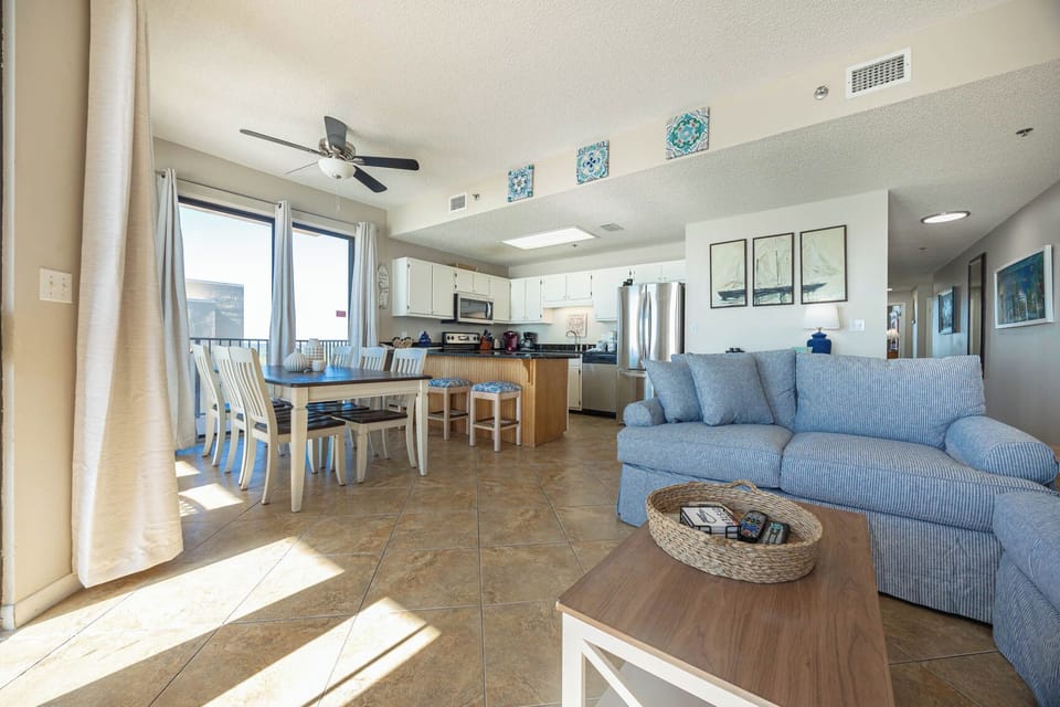 Living area featuring a blue striped sofa, dining table, and a modern kitchen with bar seating, all bathed in natural light