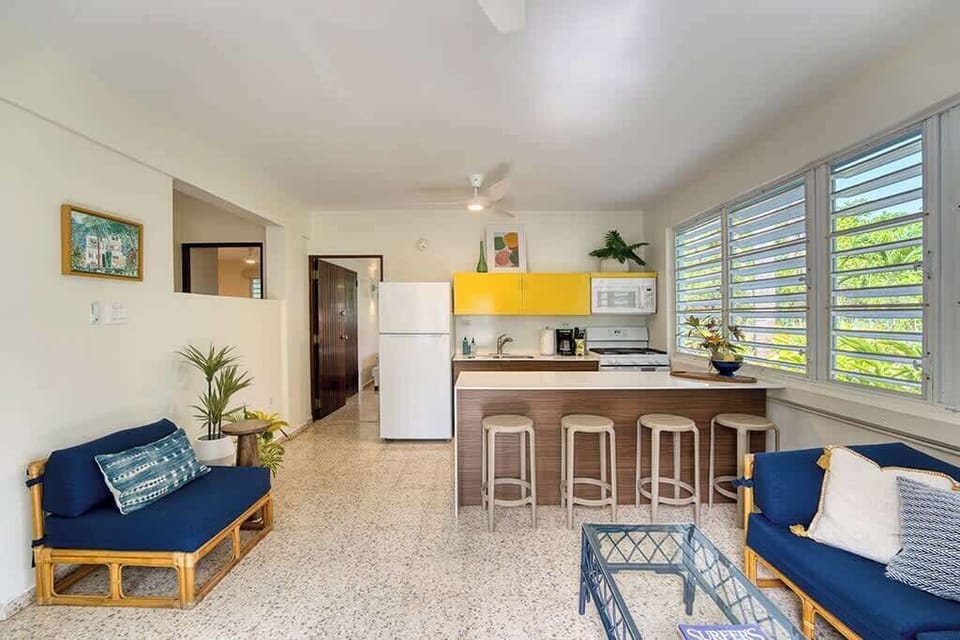 Living room seating with a smart TV and kitchen.  Overhead fan.