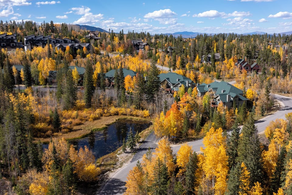Stunning aerial view of mountain community nestled among vibrant autumn foliage and evergreen forests, with distant peaks completing the scenic landscape.