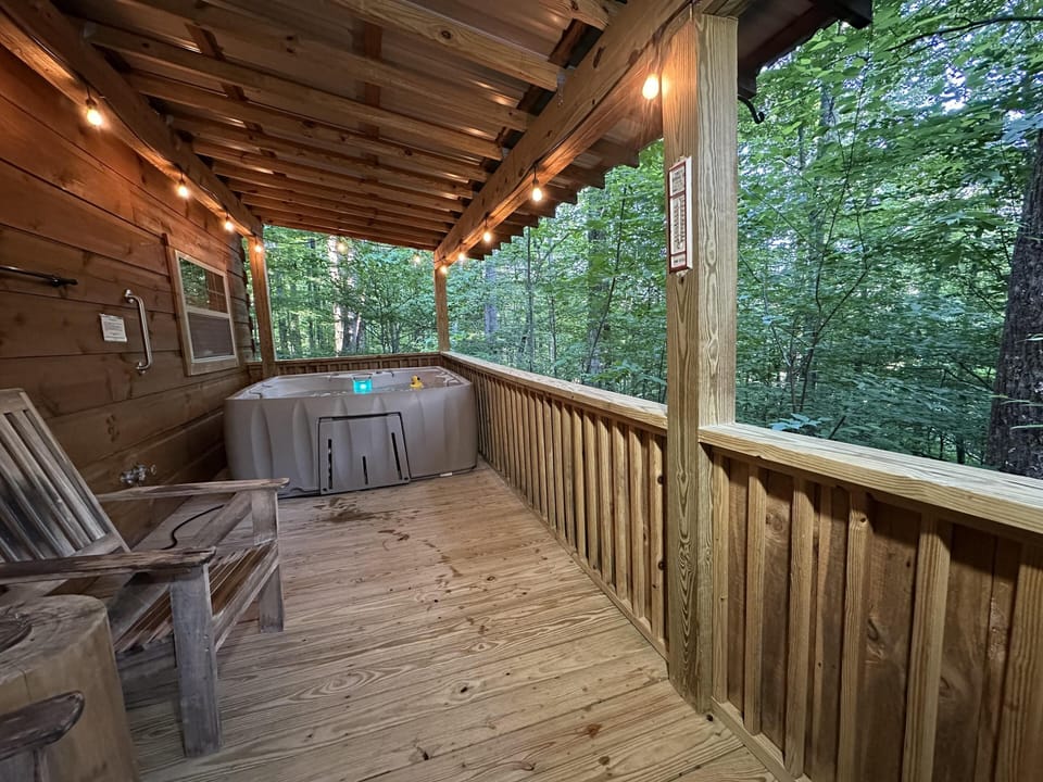 Covered wooden porch with string lights, Adirondack chair, and large hot tub overlooking dense forest.