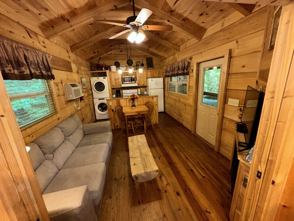 Living area with gray sofa, rustic wood coffee table, vaulted pine ceiling, and stacked washer-dryer near kitchenette.