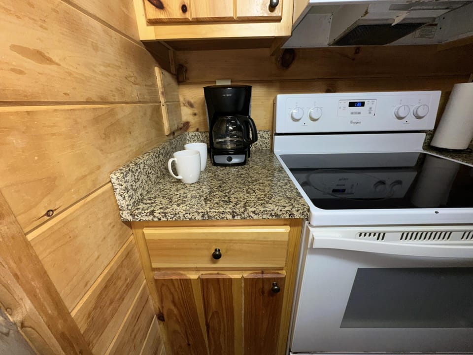 Black coffee maker sits on granite countertop next to white electric stove with wood-paneled walls.