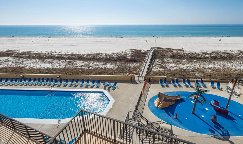 The view showcases a sparkling pool area with lounge chairs, pirate ship splash park, and a pristine beach just beyond