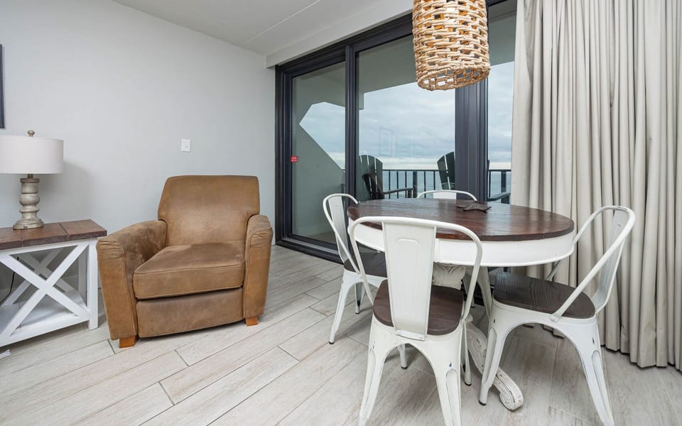 The living room features a comfortable chair and a dining table, with stunning beach views through large sliding glass doors