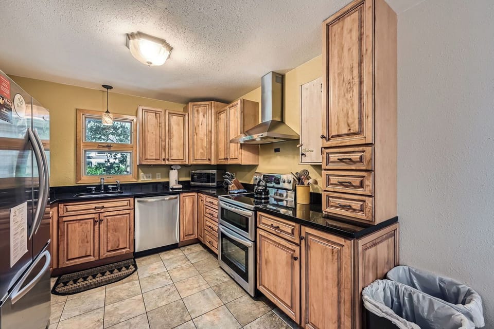 A kitchen with wooden cabinets, stainless steel appliances, including a refrigerator, stove, and dishwasher, a small window above the sink, and tiled floors. A trash bin is visible in the corner.