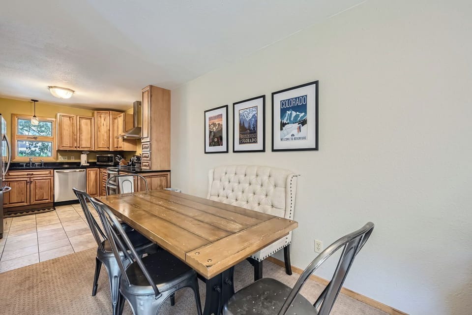 Dining area with a wooden table, metal chairs, and a tufted bench. Kitchen in the background with wooden cabinets and stainless steel appliances. Three framed posters on the wall.