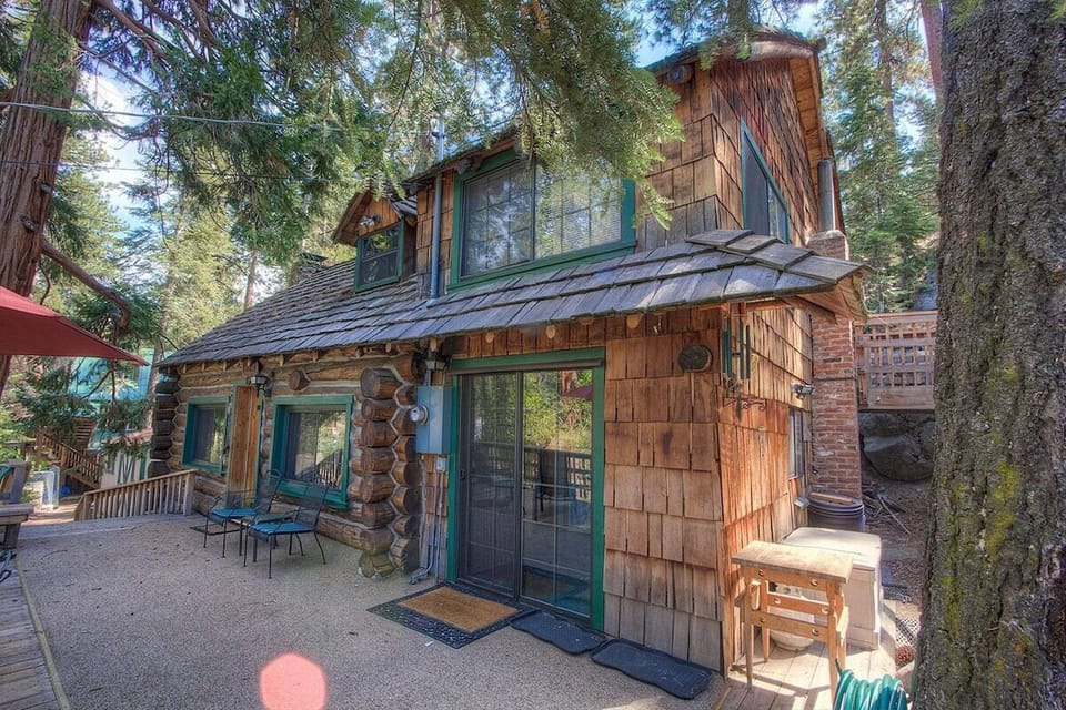 Cabin exterior with sliding door, windows, patio, patio chairs, umbrella, stairs, tree, and hose.