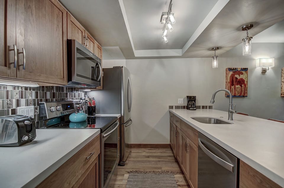 Modern kitchen with stainless steel appliances, wooden cabinets, a tiled backsplash, and quartz countertops. The room features a sink island and overhead lighting with a dining area visible in the background.