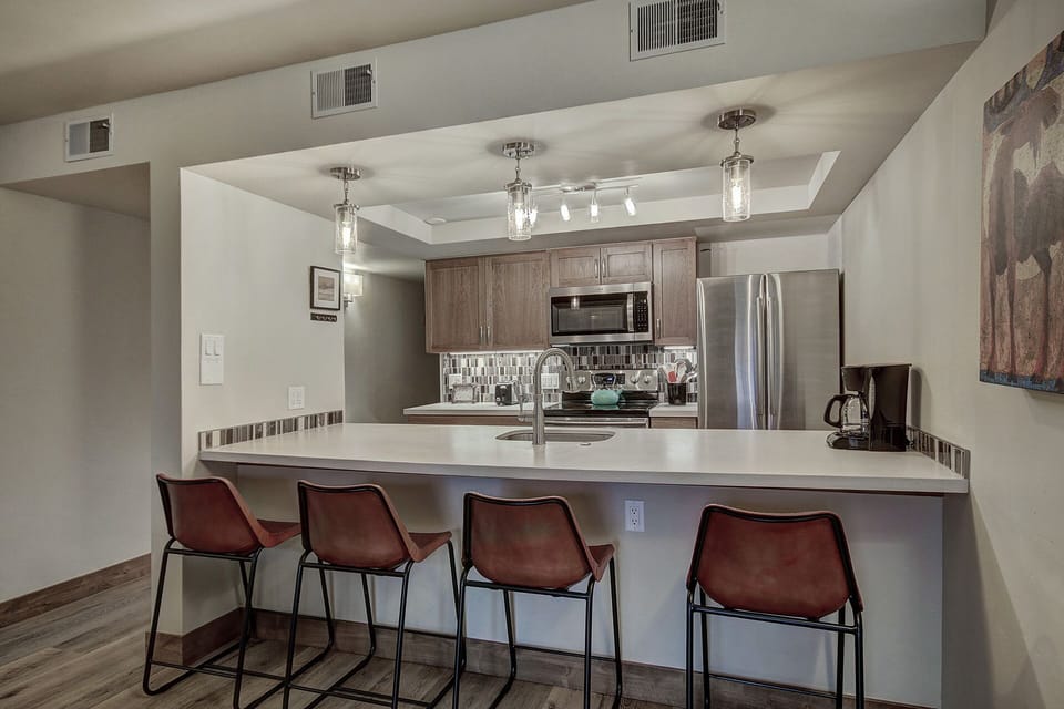 A modern kitchen with a central island, four bar stools, stainless steel appliances, and wooden cabinetry. The ceiling has recessed lighting and pendant lights hang above the island.