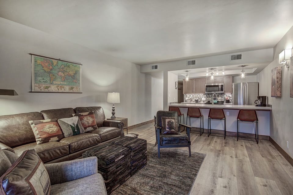 A modern living area with a brown leather sofa, chairs, a wall-mounted map, a lamp, a rug, a kitchen island with four stools, and a kitchen with wooden cabinets and stainless steel appliances.