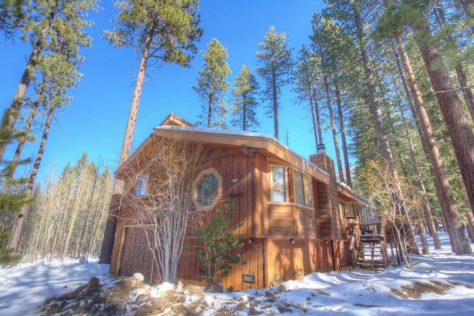 Snow-season exterior with garage door, windows, chimney, deck stairs, snow, and trees.