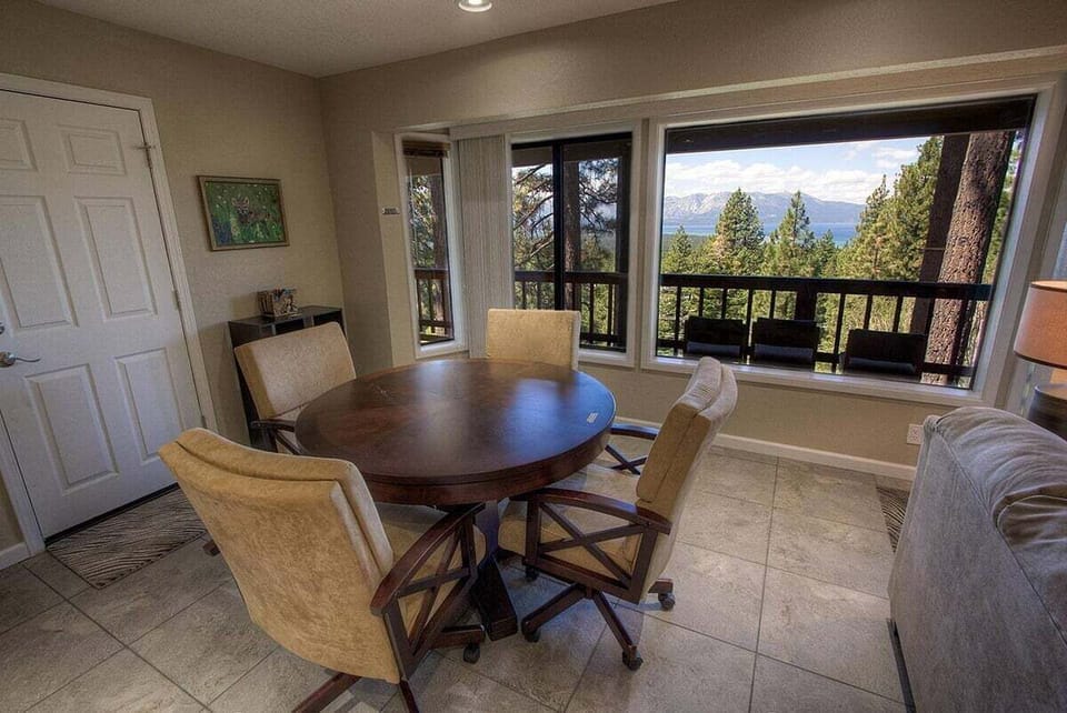Game area with game table, chairs, windows, balcony railing, lake view, mountains, and trees.