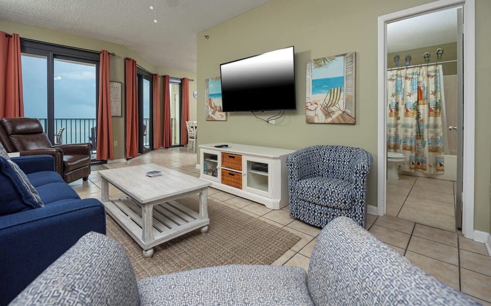 The living area features a sofa with queen sleeper, wall-mounted TV, and large windows offering a view of the beach