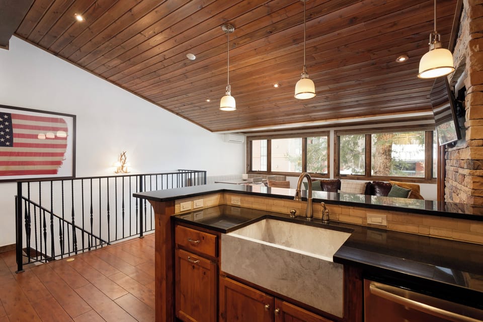 A kitchen with wooden ceiling and floor, a marble sink, and hanging pendant lights. A window with a view of trees is in the background, and a staircase with a black metal railing is on the left.