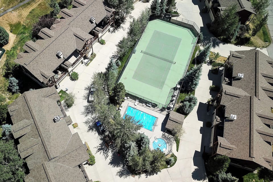 Aerial view of residential complex featuring tennis court, swimming pool, and hot tub surrounded by mature trees and well-maintained grounds.