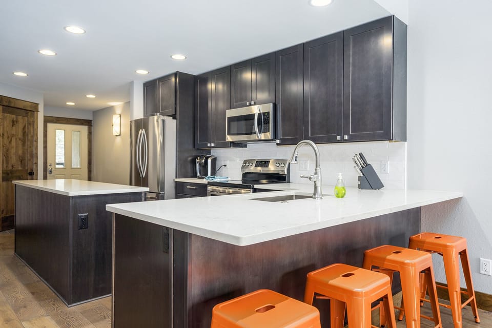Modern kitchen with dark wood cabinets, stainless steel appliances, and white countertops. An island features a sink and three orange stools, illuminated by recessed lighting.