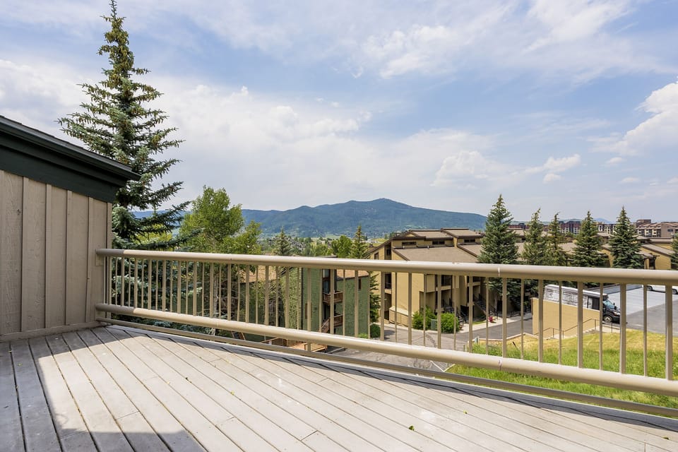 A wooden deck with railing overlooks a neighborhood of buildings set against a backdrop of green hills and a partly cloudy sky.