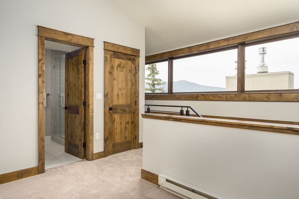 A small upstairs landing with carpeted floor, two wooden doors, and a window overlooking a mountain. One of the doors leads to a bathroom with tiled walls.