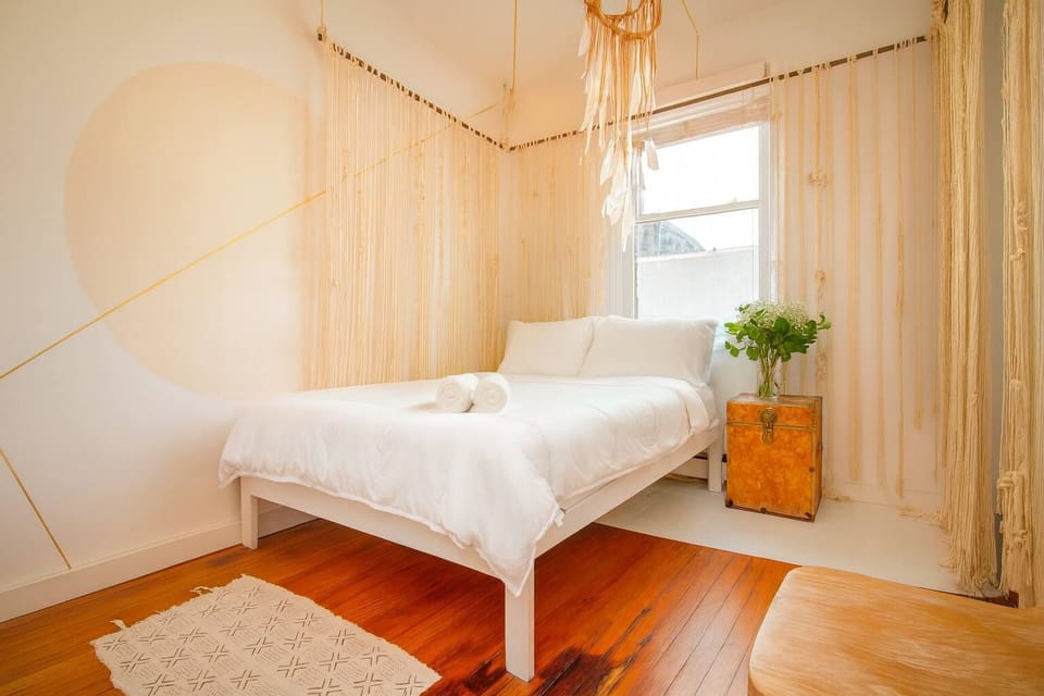 Bright, serene bedroom with airy curtains, cozy white linens, and warm wood floors—clean, calming, and beautifully styled.

