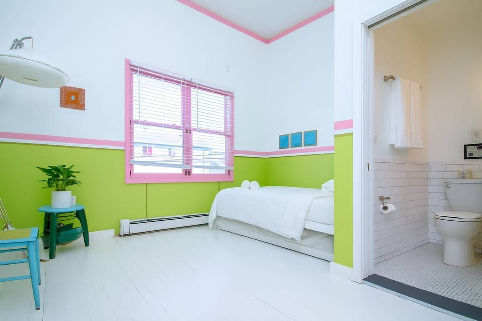 Bright, colorful bedroom with cozy bed, green accents, and ensuite bathroom—sunlit, fresh, and thoughtfully designed for comfort.
