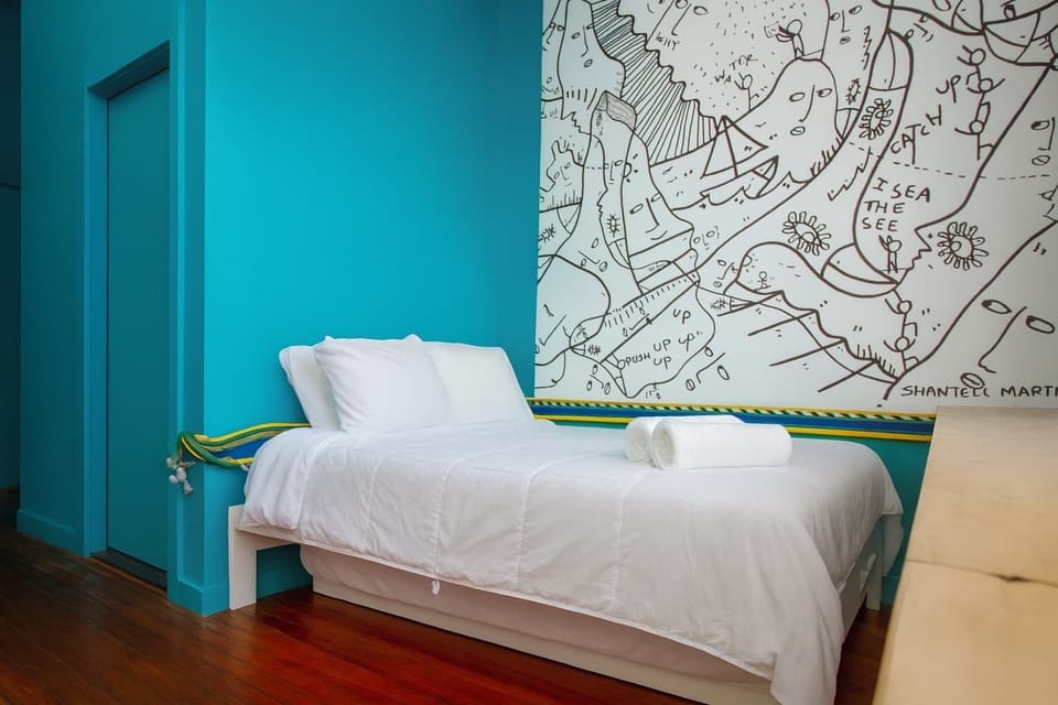 Bright, modern bedroom with bold teal walls, custom art, and crisp white linens—clean, stylish, and full of character.
