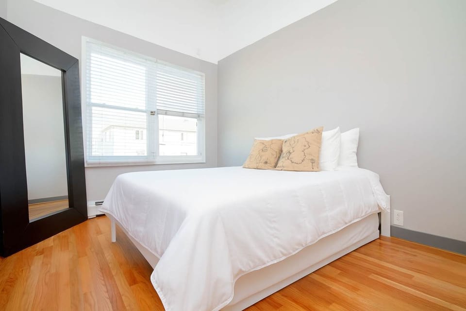 Bright, minimalist bedroom with hardwood floors, sunlit windows, and a cozy bed dressed in fresh white linens for a restful stay.
