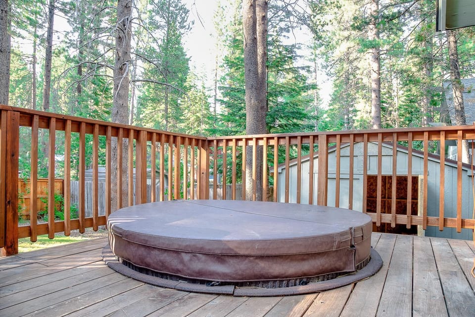 Wooden deck features a covered hot tub surrounded by pine trees and a gray storage shed in the backyard.