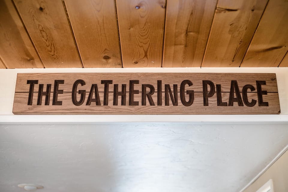 Wooden sign with "The Gathering Place" carved in bold letters mounted below a natural wood plank ceiling.