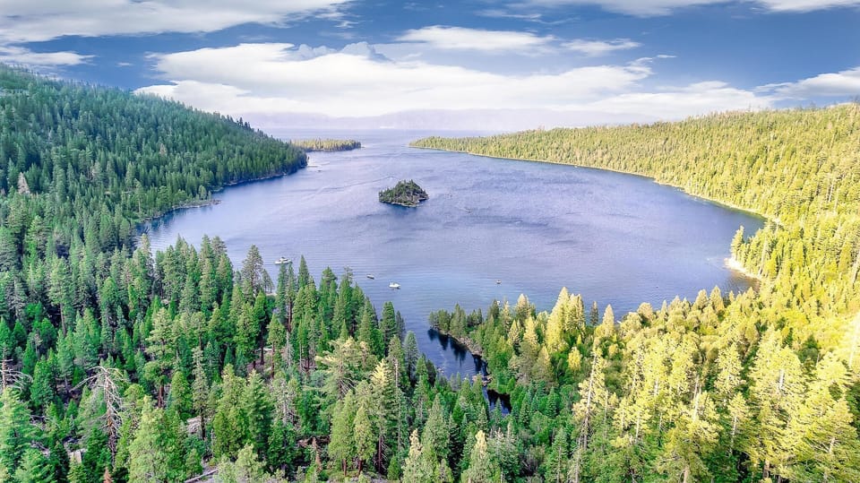 Aerial view of Lake Tahoe surrounded by dense pine forests under a bright blue sky with scattered clouds.