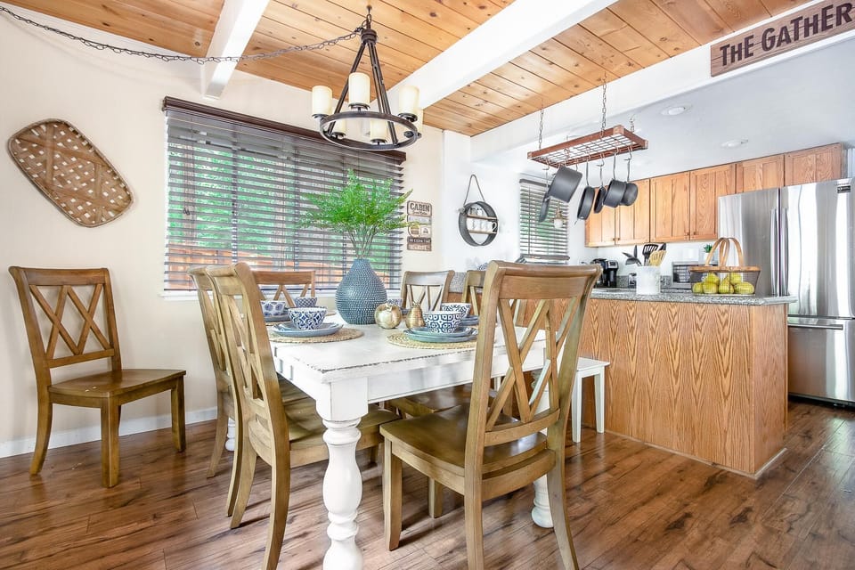Dining area with a white farmhouse table, wooden cross-back chairs, and a pottery vase centerpiece beneath a wood-paneled ceiling.
