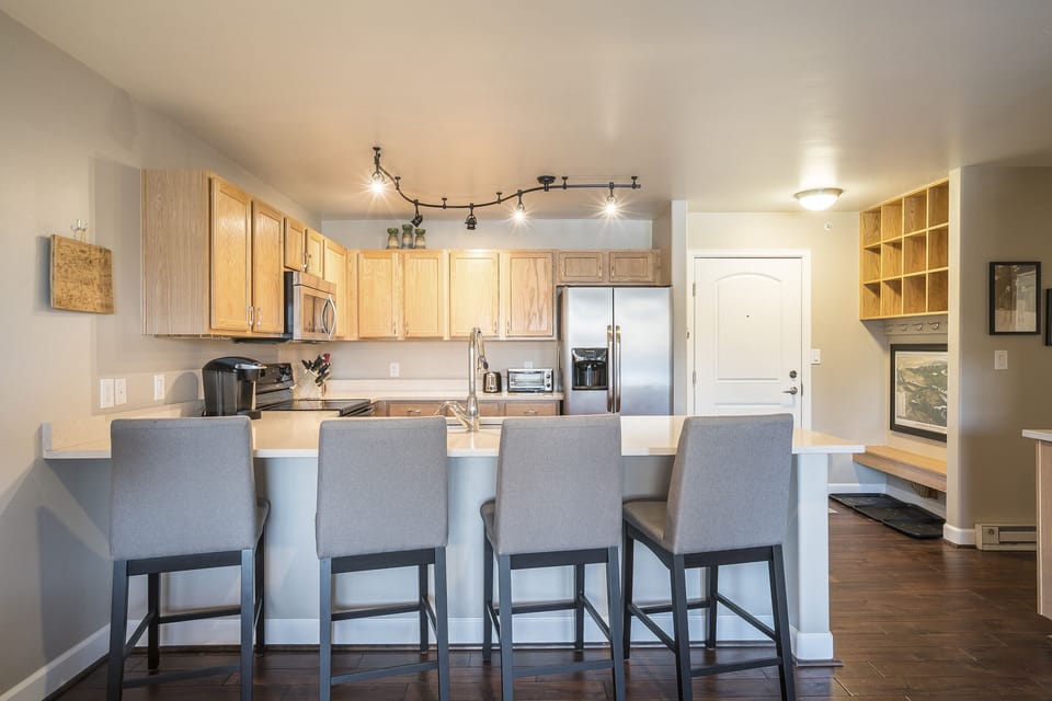 A modern kitchen with a wooden island and four gray barstools, light wood cabinets, stainless steel appliances, and a white door in the background.