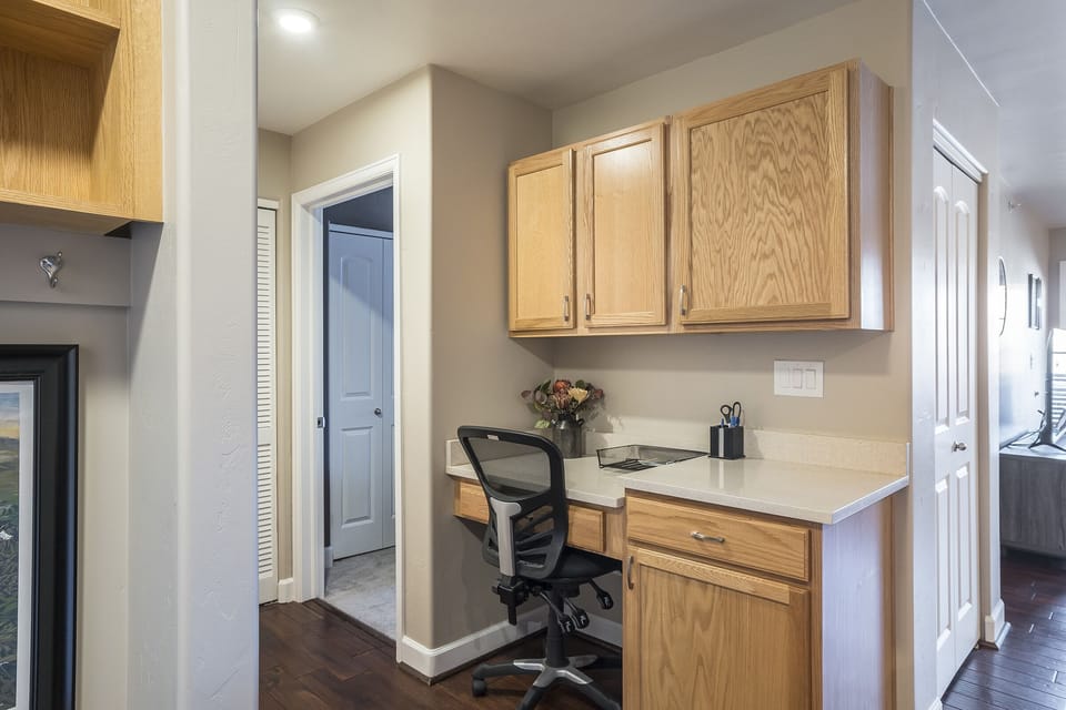 A compact home office corner with a wooden desk, a chair, a laptop, a vase with flowers, and cabinets overhead, positioned near a hallway and adjacent rooms.