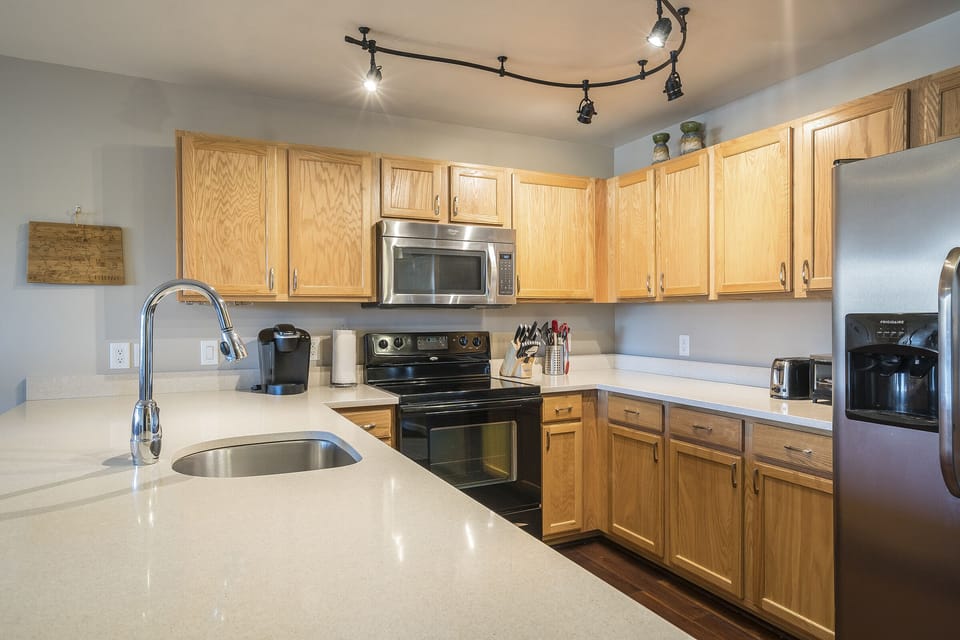 A modern kitchen with wooden cabinets, stainless steel appliances including a microwave and refrigerator, light countertops, and a chrome faucet. Track lighting is installed on the ceiling.
