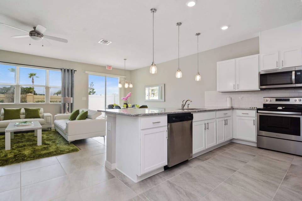 Open-concept kitchen and living area with granite island, stainless appliances, white leather sectional and green accents; sliding patio doors fill the space with sunlight.