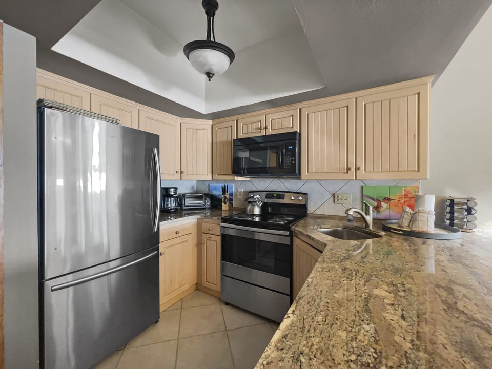 A modern kitchen with light wood cabinets, stainless steel refrigerator, stove, microwave, and granite countertops. The counter has a sink and various kitchen items.