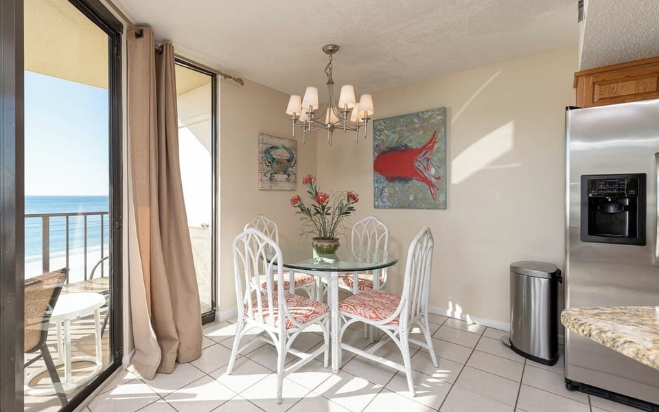 Dining area featuring a glass table with white wicker chairs, colorful artwork, and a view of the beach through large windows