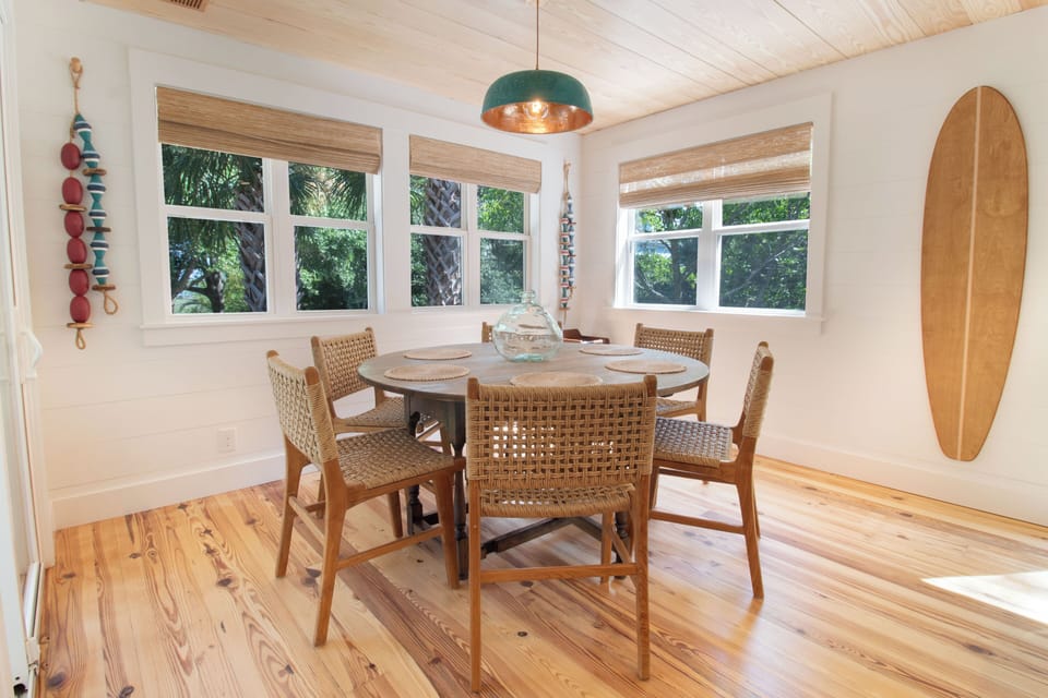 Gather around your sunny dining table with woven chairs and coastal charm—bright windows frame lush greenery, while warm wood floors and surf-inspired decor welcome easy island mornings.